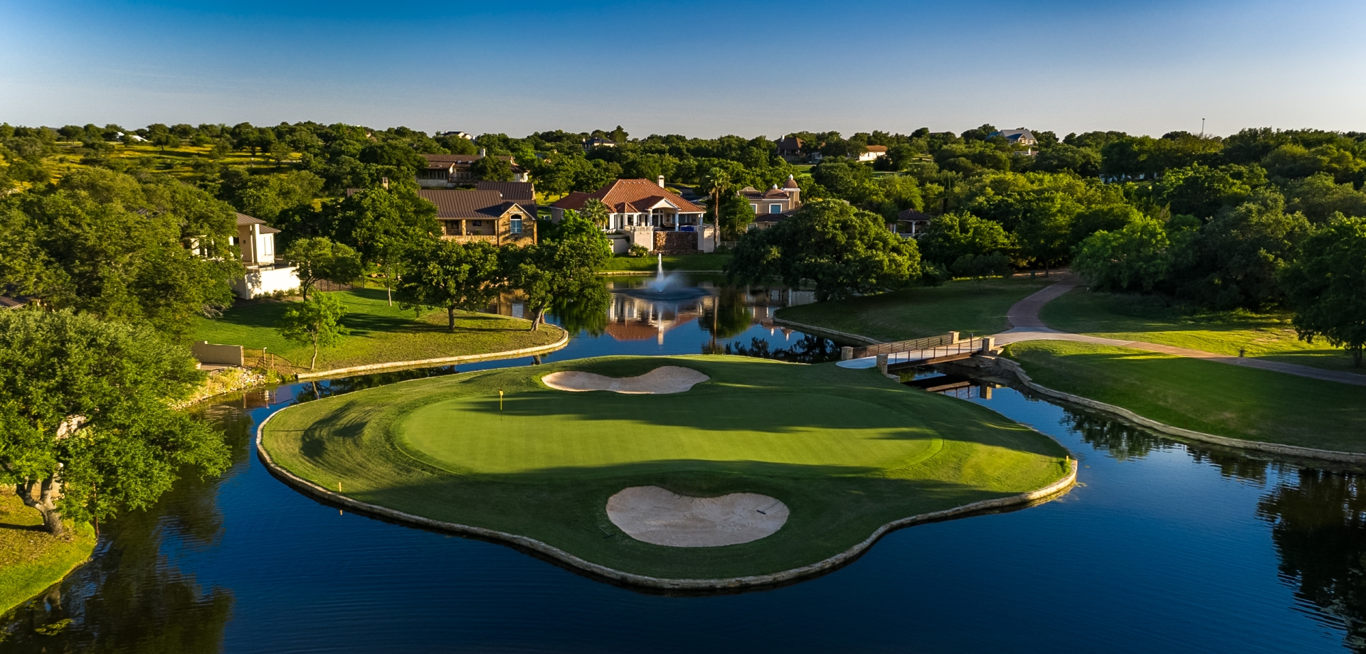 A golf green completely surrounded by water with homes in the background.